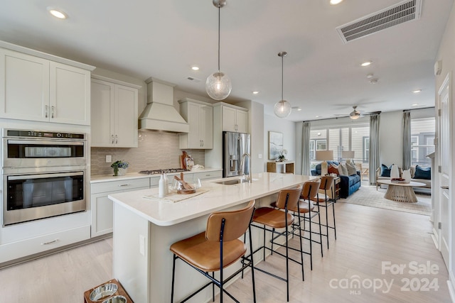 kitchen featuring appliances with stainless steel finishes, custom exhaust hood, light wood-type flooring, a kitchen island with sink, and pendant lighting