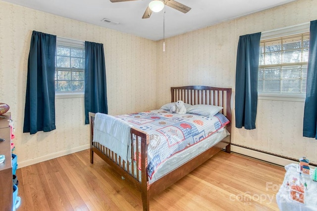 bedroom with ceiling fan, a baseboard heating unit, wood-type flooring, and multiple windows