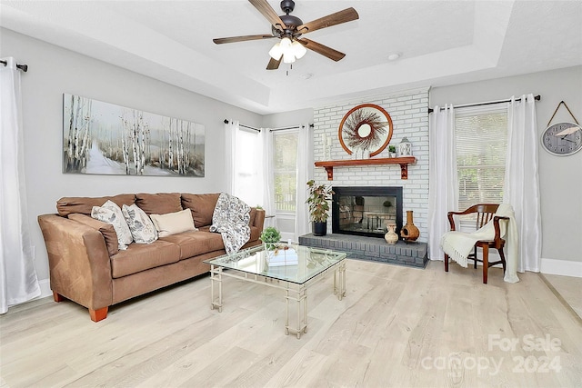 living room with light wood-type flooring, a tray ceiling, a brick fireplace, and ceiling fan