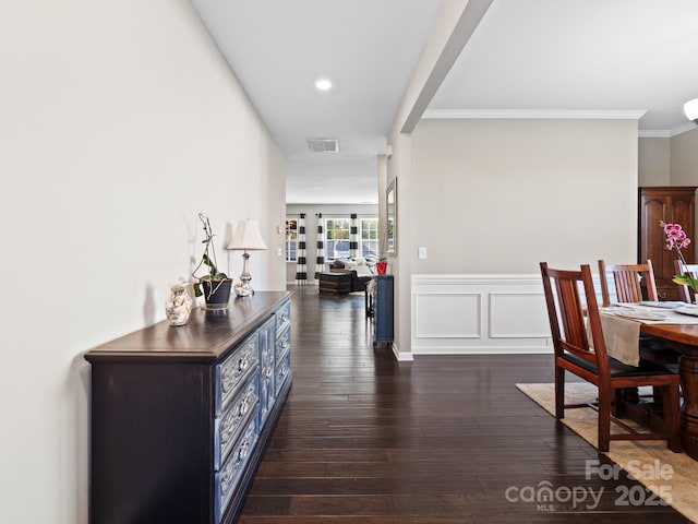 hallway featuring dark wood-style flooring, crown molding, visible vents, a decorative wall, and wainscoting