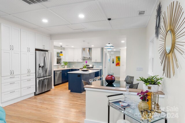 kitchen with appliances with stainless steel finishes, wall chimney range hood, pendant lighting, white cabinets, and a kitchen island