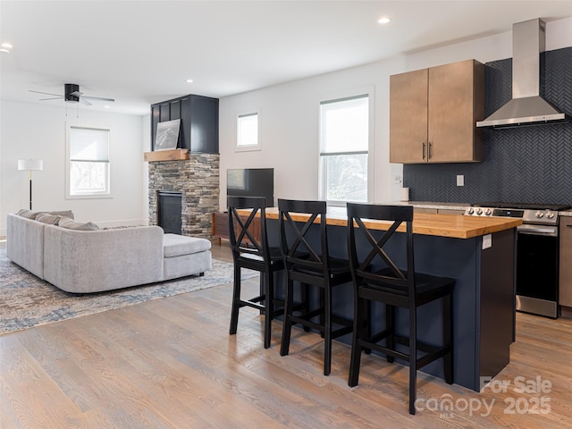 kitchen with a fireplace, open floor plan, stainless steel range oven, wall chimney range hood, and a kitchen bar