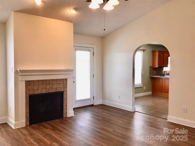 unfurnished living room with dark wood-style floors, a tile fireplace, arched walkways, and baseboards