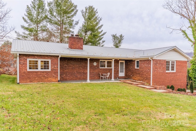 rear view of property featuring a patio, a yard, a chimney, brick siding, and metal roof