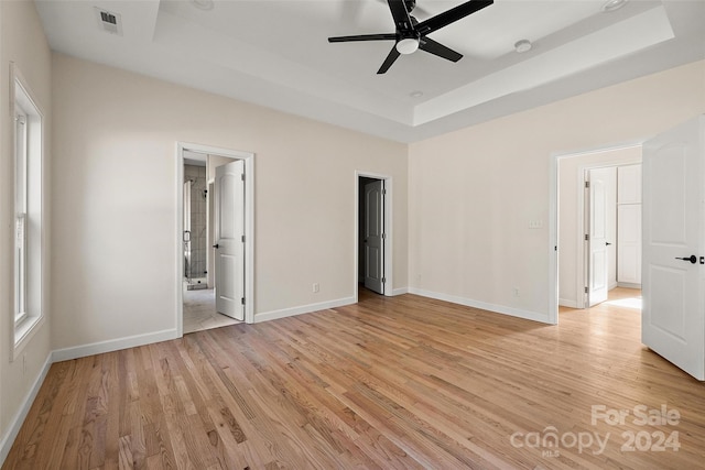 unfurnished bedroom featuring a spacious closet, ensuite bath, light wood-type flooring, a raised ceiling, and ceiling fan