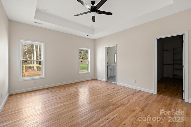 unfurnished bedroom featuring ensuite bath, a tray ceiling, a spacious closet, and light wood-type flooring