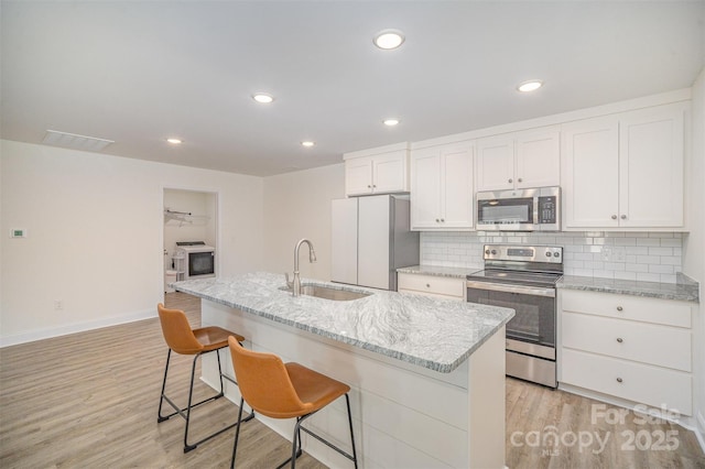 kitchen with light stone counters, a kitchen island with sink, stainless steel appliances, white cabinetry, and a sink