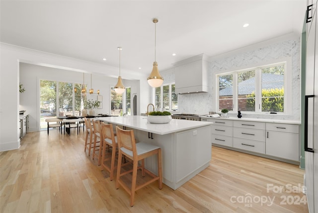 kitchen with a center island with sink, white cabinets, light hardwood / wood-style flooring, decorative light fixtures, and custom range hood