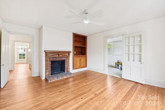 unfurnished living room with built in shelves, light hardwood / wood-style flooring, ceiling fan with notable chandelier, and a brick fireplace