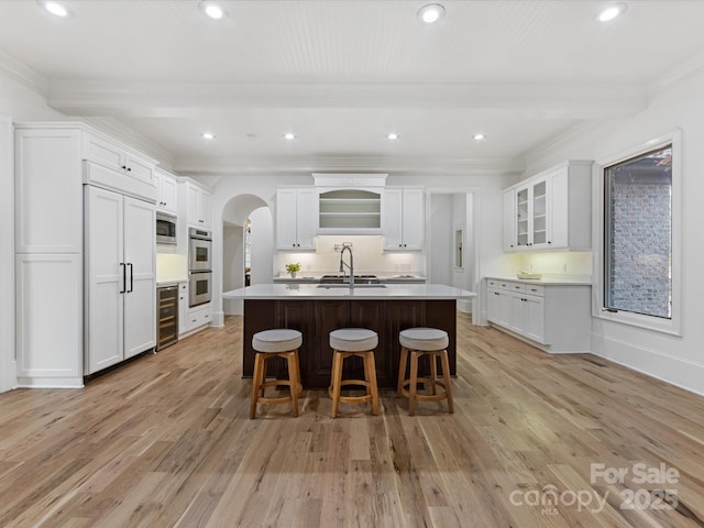 kitchen with beamed ceiling, white cabinets, beverage cooler, a kitchen island with sink, and light wood-type flooring