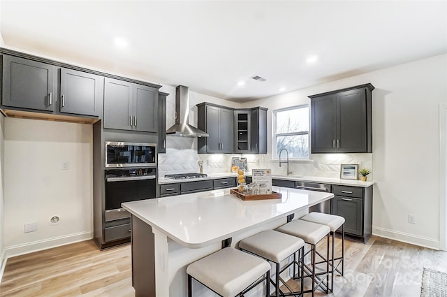 kitchen with wall chimney exhaust hood, light hardwood / wood-style floors, a breakfast bar, a kitchen island, and appliances with stainless steel finishes