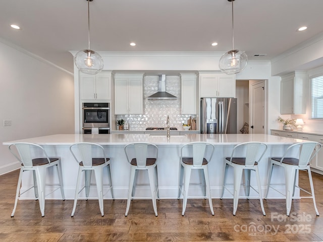 kitchen featuring a large island with sink, decorative light fixtures, wall chimney range hood, and appliances with stainless steel finishes