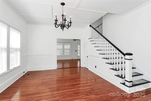 staircase featuring a wainscoted wall, crown molding, wood finished floors, ceiling fan with notable chandelier, and visible vents