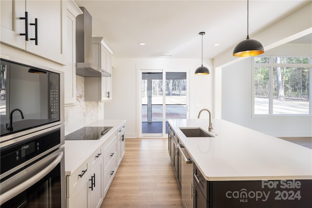 kitchen featuring white cabinets, an island with sink, black appliances, and wall chimney range hood