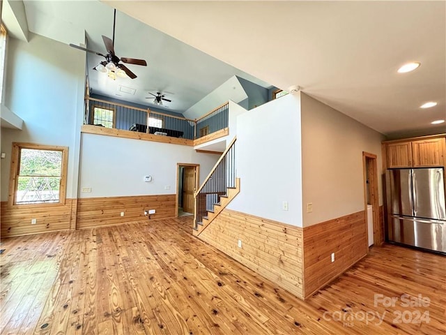 unfurnished living room with ceiling fan, a towering ceiling, wooden walls, and light hardwood / wood-style flooring