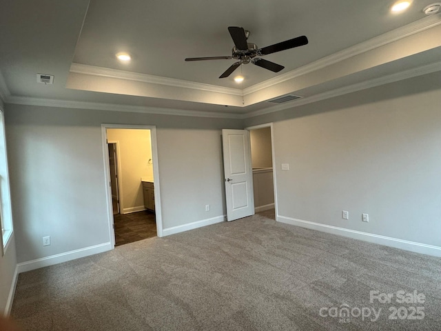 unfurnished bedroom featuring ensuite bath, ornamental molding, a tray ceiling, dark carpet, and ceiling fan