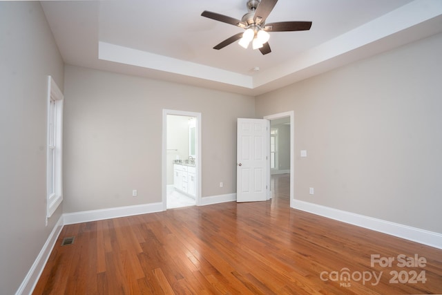 unfurnished bedroom featuring wood-type flooring, connected bathroom, a tray ceiling, and ceiling fan