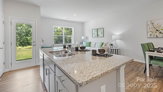 kitchen with a kitchen island with sink, a sink, baseboards, light wood-style floors, and light stone countertops