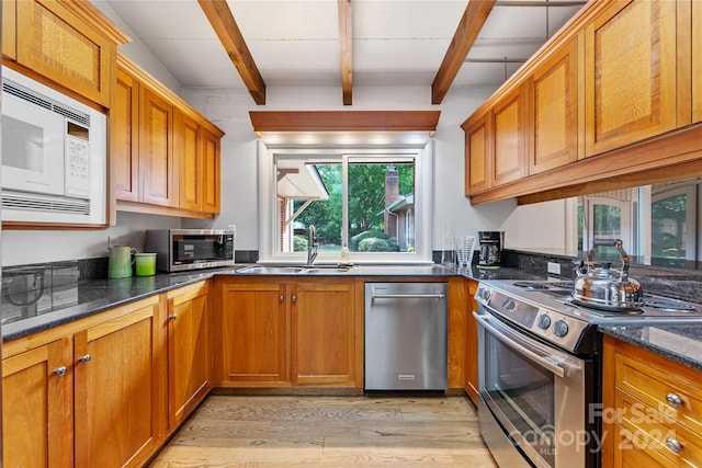 kitchen with appliances with stainless steel finishes, dark stone counters, sink, beam ceiling, and light hardwood / wood-style floors