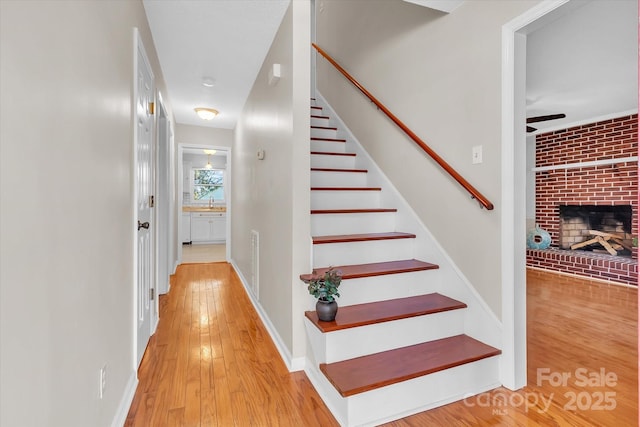 staircase with a brick fireplace, ceiling fan, visible vents, baseboards, and hardwood / wood-style floors