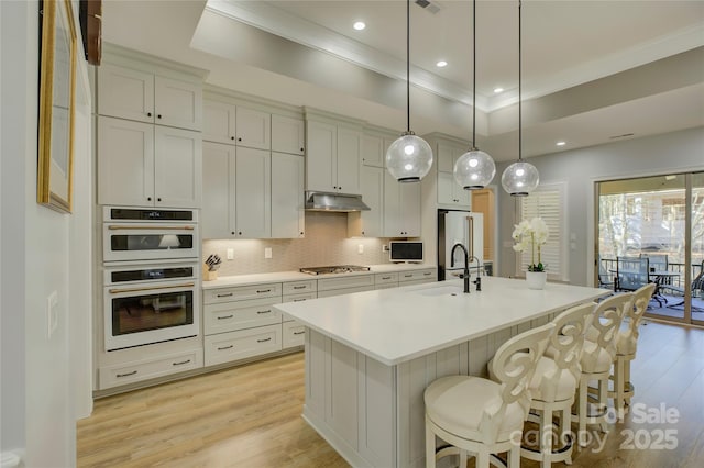 kitchen with decorative light fixtures, stainless steel appliances, a raised ceiling, and a kitchen island with sink