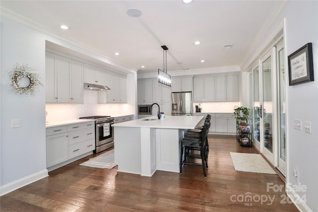 kitchen with ornamental molding, stainless steel appliances, dark wood-type flooring, hanging light fixtures, and an island with sink