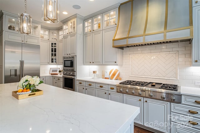 kitchen with backsplash, premium range hood, hanging light fixtures, built in appliances, and light stone counters