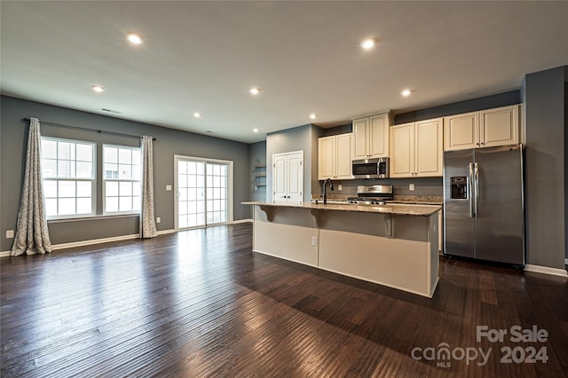 kitchen with light stone counters, stainless steel appliances, a center island with sink, dark hardwood / wood-style floors, and a breakfast bar area