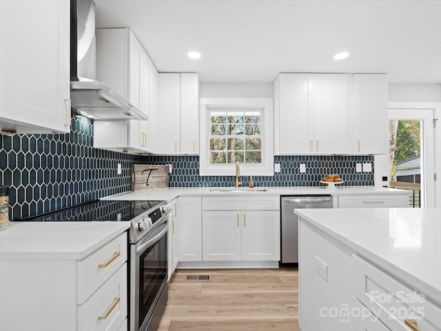 kitchen featuring white cabinets, wall chimney range hood, sink, light hardwood / wood-style flooring, and stainless steel appliances