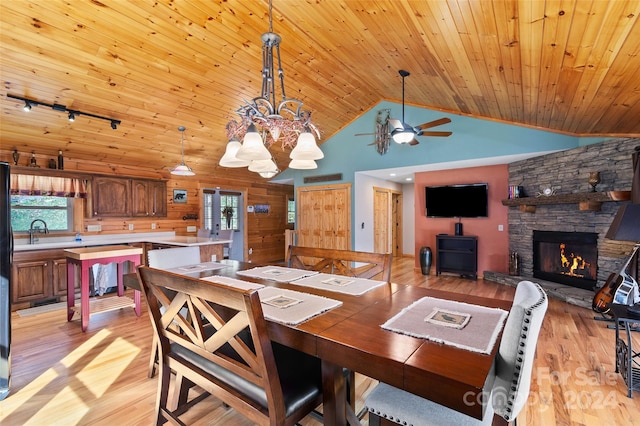 dining area with ceiling fan with notable chandelier, a stone fireplace, sink, light wood-type flooring, and wood ceiling