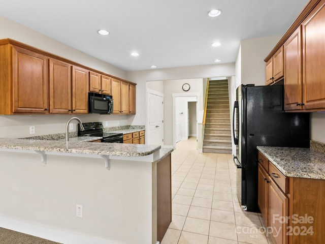 kitchen featuring kitchen peninsula, light stone countertops, black appliances, light tile patterned floors, and a breakfast bar area