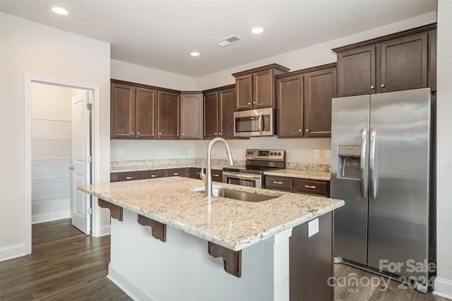 kitchen featuring a kitchen bar, appliances with stainless steel finishes, light stone counters, a center island with sink, and dark hardwood / wood-style floors