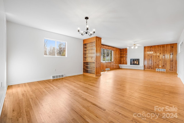 unfurnished living room with wood walls, light hardwood / wood-style flooring, ceiling fan with notable chandelier, and a brick fireplace