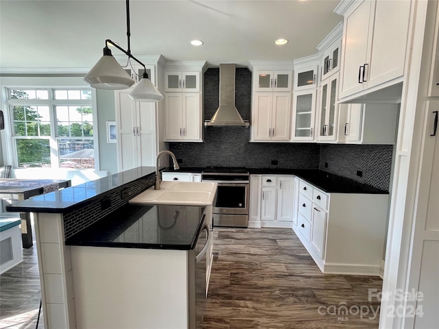 kitchen featuring stainless steel stove, hanging light fixtures, wall chimney range hood, a center island with sink, and white cabinets