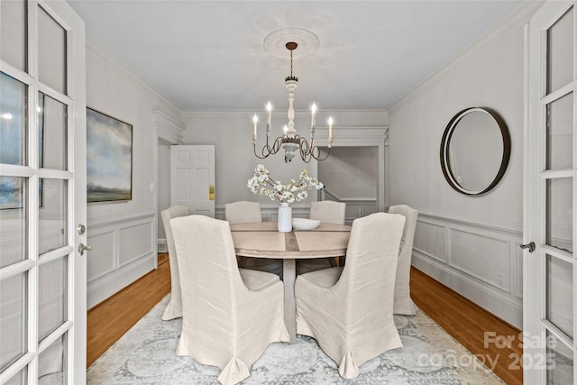 dining area with wood finished floors, a chandelier, a decorative wall, crown molding, and wainscoting