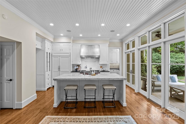 kitchen featuring wall chimney exhaust hood, white cabinets, crown molding, tasteful backsplash, and a kitchen island with sink
