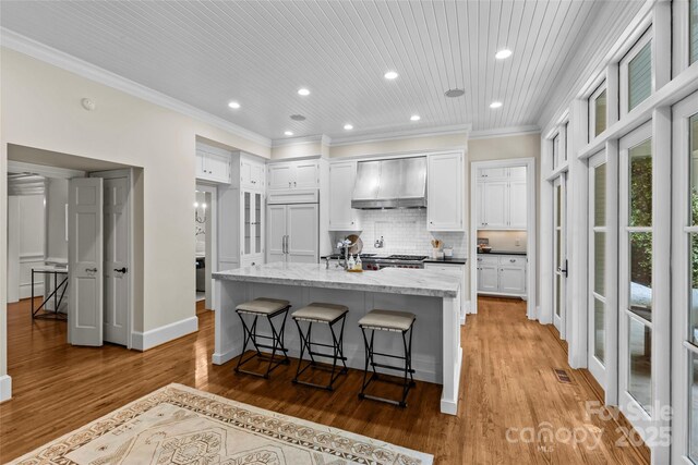kitchen featuring wall chimney range hood, recessed lighting, white cabinets, light wood-style flooring, and a center island with sink