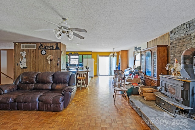 living room with ceiling fan with notable chandelier, a wood stove, wooden walls, and light parquet floors