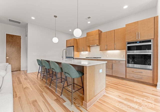 kitchen featuring stainless steel double oven, hanging light fixtures, stovetop, light hardwood / wood-style floors, and a center island with sink