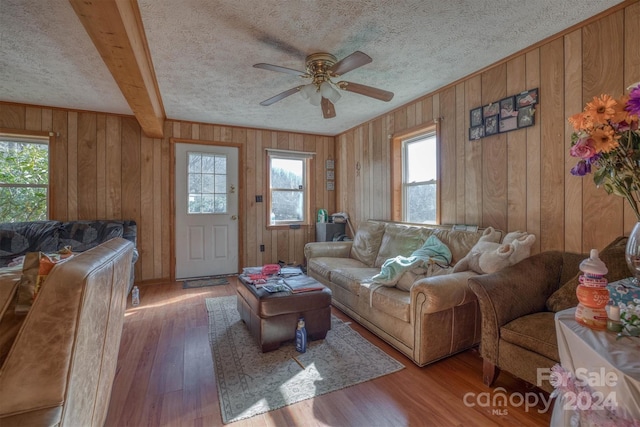 living room with wooden walls, light hardwood / wood-style flooring, ceiling fan, a textured ceiling, and beam ceiling