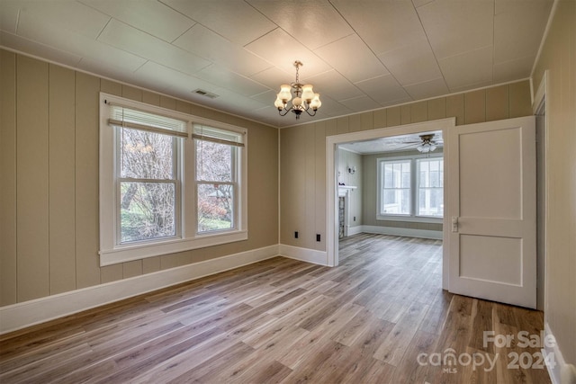 unfurnished dining area with ceiling fan with notable chandelier, light wood-type flooring, and wooden walls