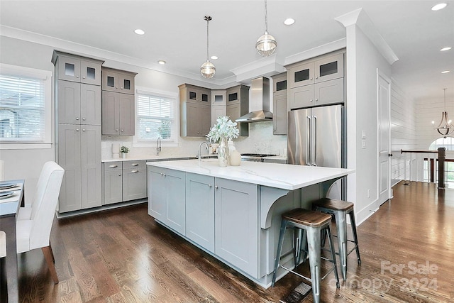 kitchen with high end refrigerator, a center island with sink, pendant lighting, and wall chimney range hood