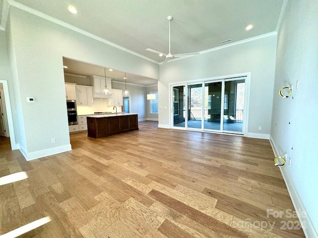 unfurnished living room with a towering ceiling, ornamental molding, ceiling fan, sink, and light hardwood / wood-style flooring