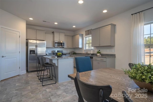 kitchen with light stone countertops, appliances with stainless steel finishes, a center island, a kitchen bar, and gray cabinets