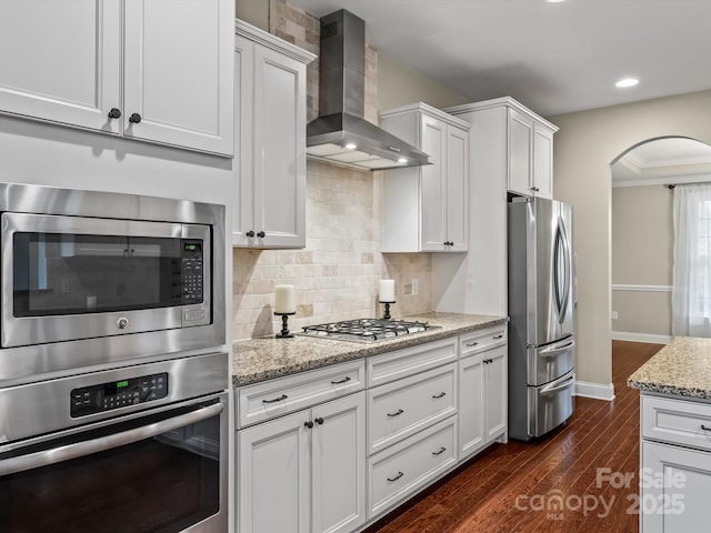 kitchen with light stone counters, wall chimney exhaust hood, stainless steel appliances, dark hardwood / wood-style flooring, and white cabinetry