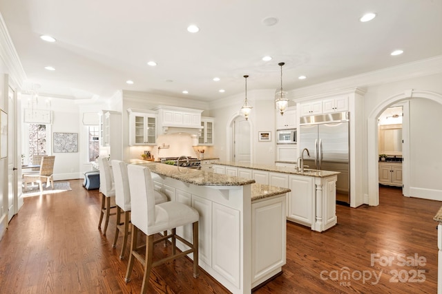 kitchen with arched walkways, built in appliances, dark wood finished floors, and a large island with sink