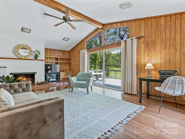 living room with a textured ceiling, ceiling fan, lofted ceiling with beams, light wood-type flooring, and a brick fireplace