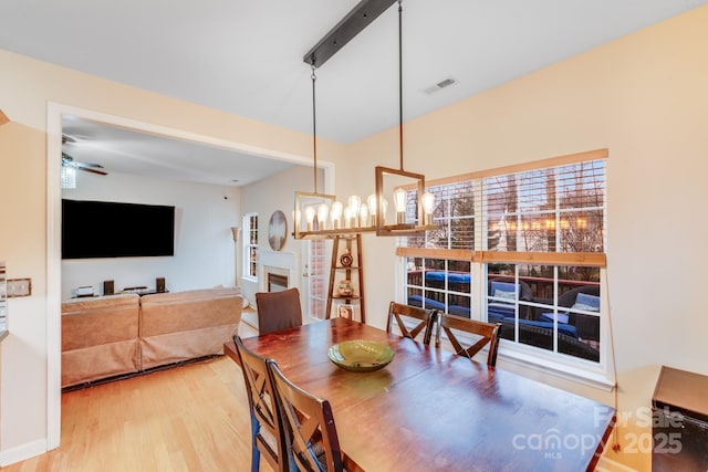 dining area with ceiling fan with notable chandelier, a fireplace, wood finished floors, visible vents, and baseboards