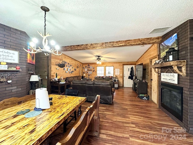 dining room with dark wood-type flooring, lofted ceiling with beams, wood walls, a fireplace, and ceiling fan with notable chandelier