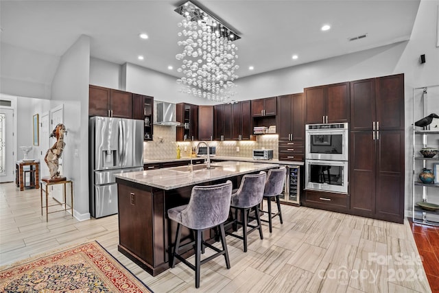 kitchen featuring a center island with sink, wall chimney range hood, sink, appliances with stainless steel finishes, and a kitchen bar